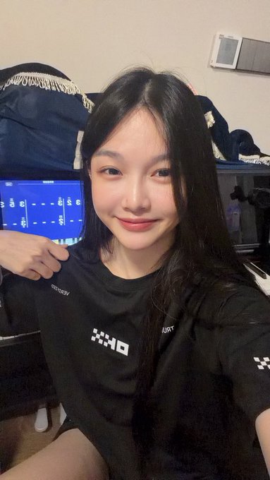 A young woman with long dark hair smiles for a selfie while wearing a black t-shirt with 'XKO' branding, with a monitor visible in the background.