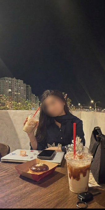 The image shows a person sitting at an outdoor table with food and drinks. The individual appears to be enjoying a casual meal outside under the lights of a cityscape at night. There are various items on the table, including two coffee cups, one closer to the top right corner and another towards the center left side, as well as two donuts - one slightly more central in the image and another further down towards the bottom left. Additionally, there is an open laptop next to a cell phone at the center of the scene. The overall atmosphere seems relaxed, with the city lights providing an urban backdrop for this outdoor dining experience.