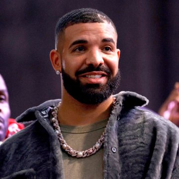 A portrait of rapper Drake smiling, wearing a gray fuzzy hooded jacket and a chain necklace.