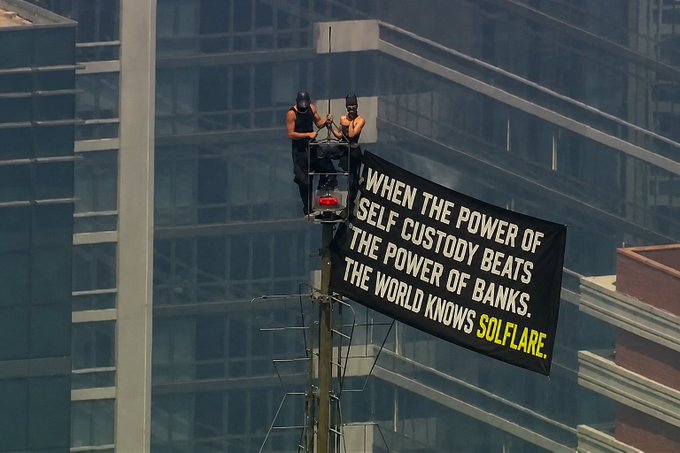 Two people sit on a billboard with a banner reading, "WHEN THE POWER OF SELF CUSTODY BEATS THE POWER OF BANKS. THE WORLD KNOWS SOLFLARE." The accompanying post "what free will looks like:" mirrors the bold act of scaling the structure to spread a decentralized finance message promoting financial autonomy.