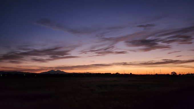 A scenic landscape photograph capturing a vibrant sunset over a darkened valley, with silhouettes of mountains and power lines set against a sky painted in deep blues, purples, and warm orange hues.