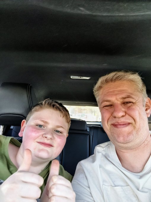 A selfie taken inside a car shows a blonde man and a young boy, both smiling and giving thumbs-up gestures to the camera.