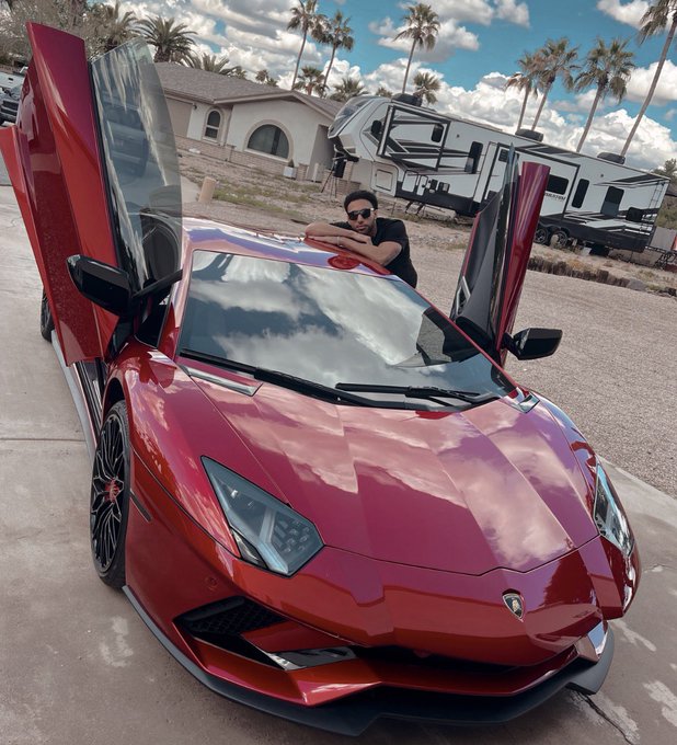 A man leans out of the windshield of a red Lamborghini with both scissor doors open, parked in a lot with an RV and palm trees in the background.