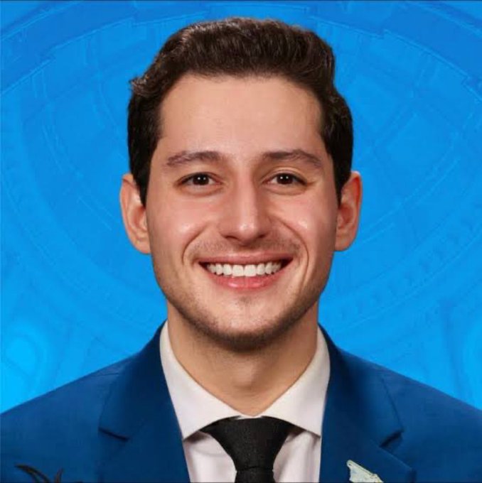 A professional headshot of a smiling young man with dark hair, wearing a blue suit, white shirt, and black tie, set against a blue patterned background.