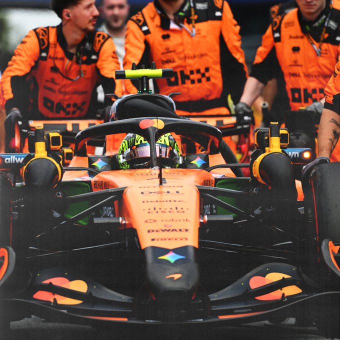 A McLaren Formula 1 car, featuring prominent sponsor logos including Dell, Cisco, DeWalt, and Richard Mille, is pushed through the pit lane by a crew wearing matching orange "OKX" uniforms, with the driver suited up in the cockpit—capturing the team in full preparation mode ahead of the Austrian Grand Prix.