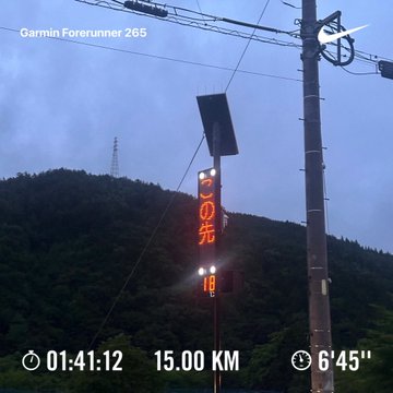 A cycling activity captured by a Garmin Forerunner 265 showing a wet road sign in Japanese with distance '15.00 KM', time '01:41:12', and pace '6'45'' against a forested hillside.