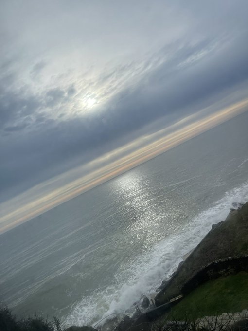 A tilted landscape photograph of an ocean view with overcast clouds, the sun breaking through, and sunlight reflecting on the water, with waves crashing against rocky cliffs in the foreground.