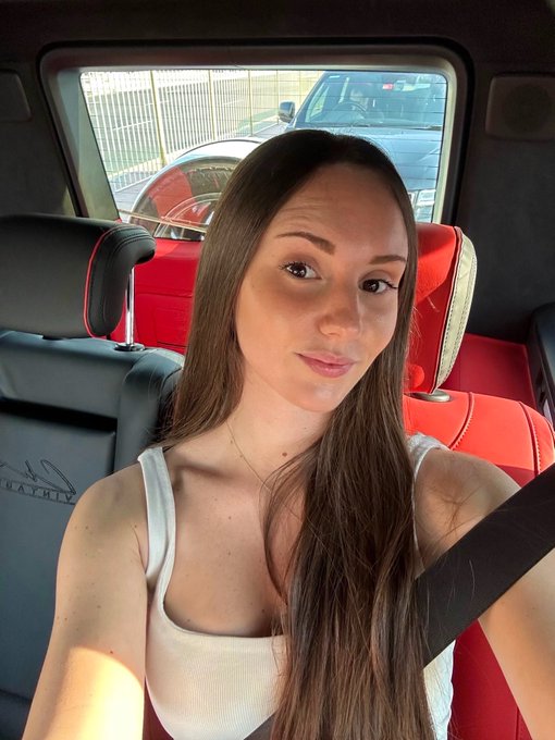 A woman with long brown hair, wearing a white tank top, takes a selfie while seated in a car with red leather interior. The caption "Happy taco Tuesday fam" doesn't directly relate to the image, as there are no tacos visible—she's simply documenting her day with a casual photo.