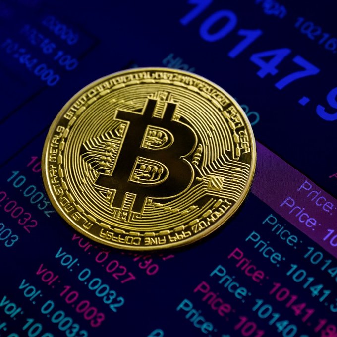 A gold Bitcoin token rests on a digital trading screen displaying a price of "10147" alongside volume data and price tickers. The image visually represents the cryptocurrency's volatility, complementing the post about Bitcoin's sharp drop below $82K and the massive wave of long-position liquidations that followed.