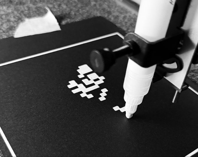 A black-and-white close-up of a robotic arm dispensing a white paste in a pixel art pattern onto a dark surface.