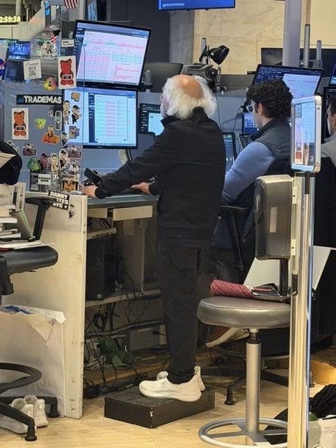 A trader wearing a 'TRADEMAS' hoodie stands at a desk with multiple monitors displaying market data, standing on a step stool while another person sits nearby.
