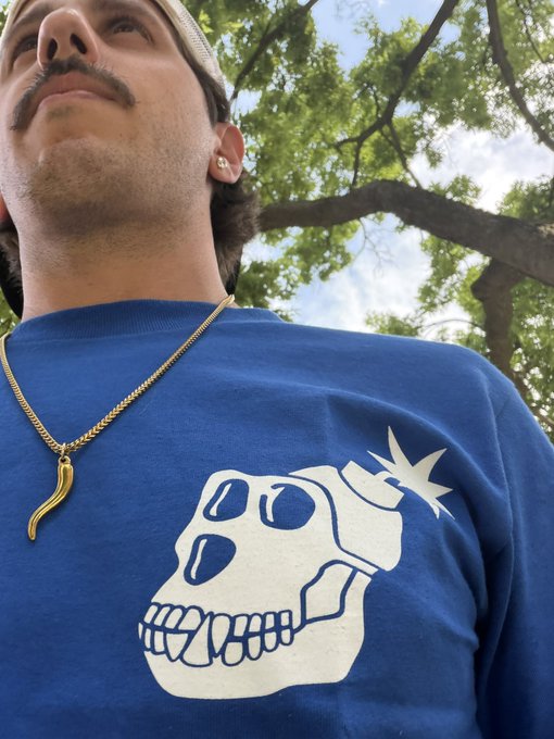 A man with a mustache and gold chili pepper pendant looks up toward the sky, wearing a blue sweatshirt featuring a white skull graphic with a lit bomb fuse. The outdoor setting with trees in the background complements the post announcing his excitement for the first Midwest Ape IRL meetup in Chicago.