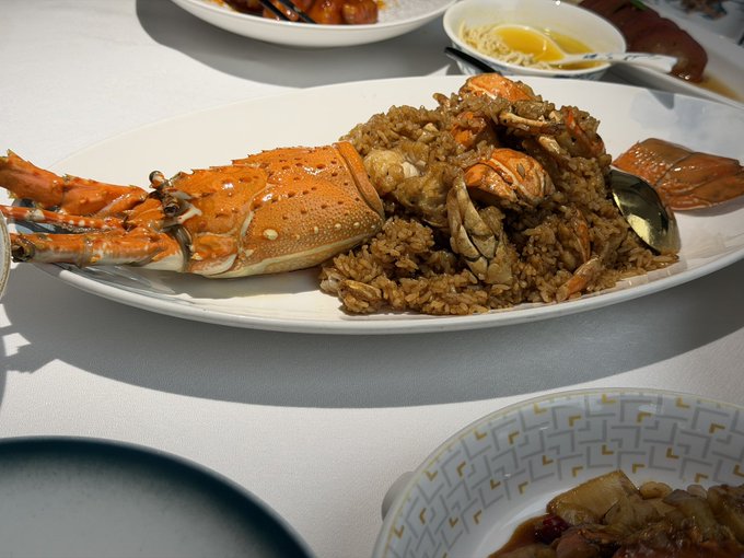 A close-up of a dining table featuring a plate of crab fried rice with large crab pieces, alongside other dishes including one with noodles and dipping sauce, and another with a dark sauce.