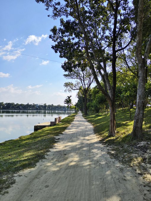 The image shows a long dirt road that runs through a park with trees lining both sides of the path, leading towards a lake. The sky above appears hazy, suggesting an early morning or late afternoon setting. In the background, there are buildings visible along the shoreline, indicating that this park is located near a city or town. A post on social media has been shared about the finish line for a race taking place in the area, and it encourages people to keep going by sharing their progress with others.

The image also contains some birds flying overhead, adding life and movement to the scene. The overall atmosphere of the photo is one of tranquility and leisure as people enjoy the park's natural beauty while participating in or watching a race.