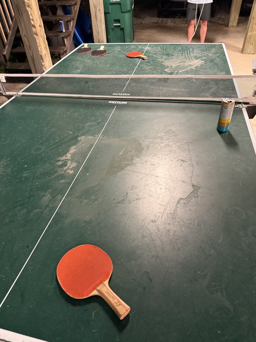A worn, green ping pong table set up outdoors, with an orange paddle and a beverage can resting on the surface.