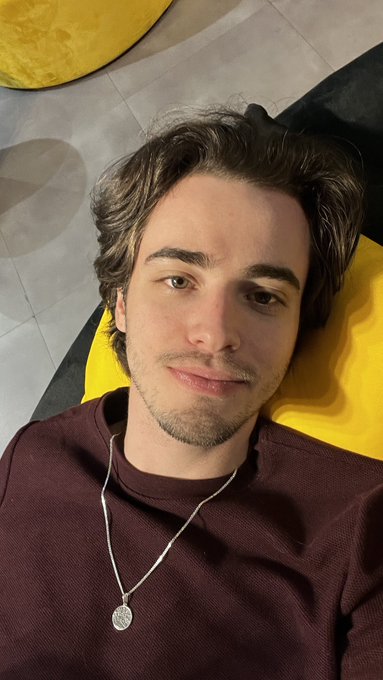 A selfie of a young man with brown hair wearing a maroon shirt and silver pendant necklace, lying on a yellow surface.
