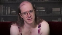 A man with long hair and glasses wearing a patterned tank top sits in front of a bookshelf, speaking to the camera.