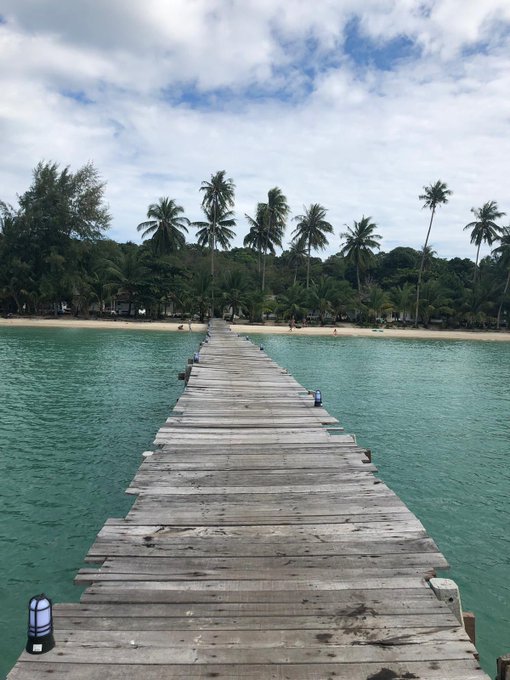 A long wooden pier extends across turquoise tropical water toward a sandy beach lined with palm trees under a partly cloudy sky.
