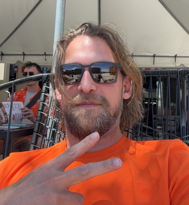 A man with long blonde hair, a beard, and dark sunglasses wears an orange shirt and holds up a peace sign at what appears to be an outdoor event under a tent.