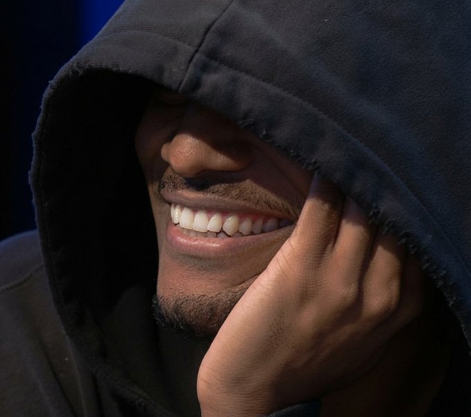 A close-up portrait of a smiling man wearing a dark hoodie pulled over his head, with his hand resting near his face.