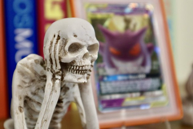 A skeleton figurine in a contemplative pose appears beside a Pokémon trading card, with partial text "365" and "5" visible on items in the background. The spooky, Halloween-themed image pairs perfectly with the post's reference to "Spooky season" and "frightfully exciting" October, using the skeleton as a playful nod to the eerie holiday spirit.