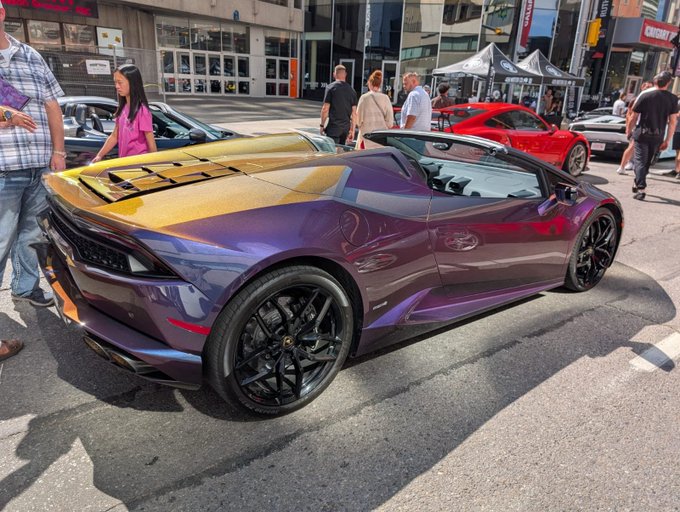 A purple-to-gold color-shifting Lamborghini Huracán Spyder parked on a city street, surrounded by pedestrians and other vehicles at what appears to be a car show.