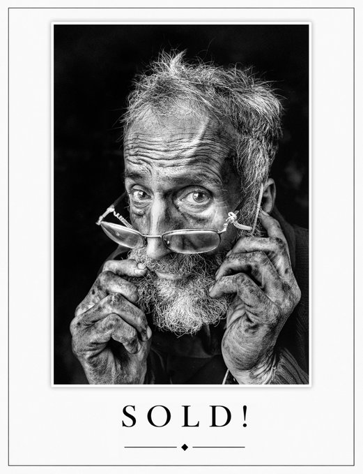 A striking black and white portrait shows an elderly, weathered man with a long beard holding his glasses to his face, captured in a powerful close-up that highlights the deep lines and texture of his hands and features. The bold text "SOLD!" displayed beneath the photograph celebrates the artist's rapid success, as the image relates to the accompanying message about an NFT collection that sold out in 10 minutes, collecting 0.888 ETH with gratitude toward collector @chidgey_david.