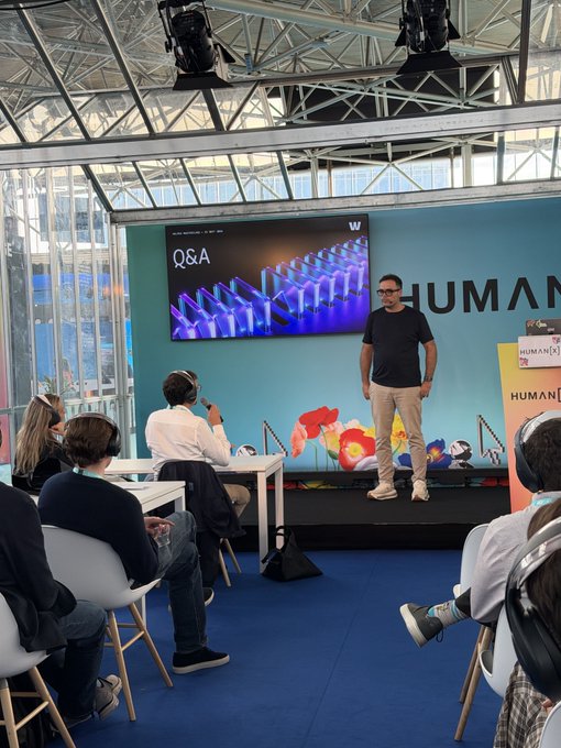 At the HumanX conference, a speaker addresses the audience during a Q&A session displayed on the large screen behind him, engaging with questions about the use of technology for personal versus corporate applications.