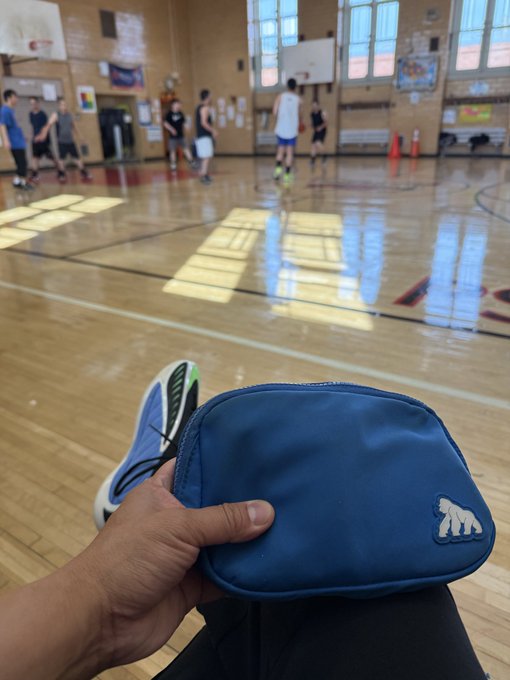 A first-person view shows a hand holding a blue zipped pouch with a white bear logo in the foreground, while a basketball game is being played on a wooden gymnasium court in the background.