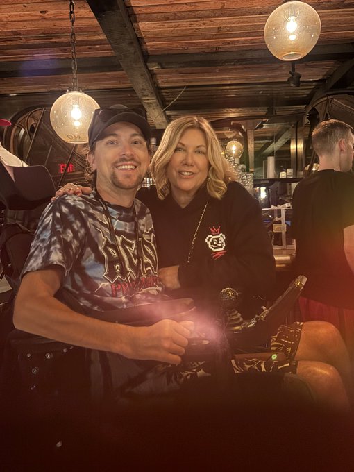 A man wearing a tie-dye t-shirt and a black cap sits next to a smiling blonde woman in a dark sweater inside a dimly lit restaurant with hanging globe lights.