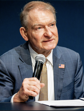 An older man in a blue checkered suit with a gold tie and American flag pin speaks into a microphone during what appears to be a panel or press event.