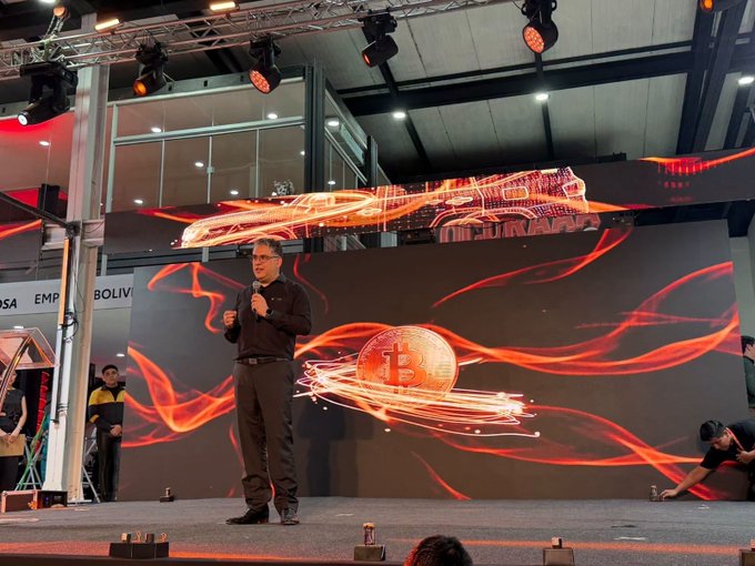 A man presents on stage at Expocruz 2026 in Bolivia, with a large digital display behind him featuring fiery orange graphics of a car and a Bitcoin symbol, illustrating the launch of BitGo-powered Bitcoin payment options for purchasing Toyota vehicles through TOYOSA. Visible banners reference "Bolivia," tying the scene to the milestone announcement of accepting Bitcoin alongside bolivianos, USD, and USDT for automotive purchases.