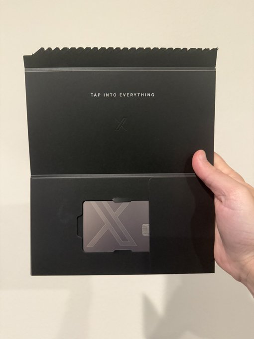 A hand holds open a sleek black XMoney packaging envelope with "TAP INTO EVERYTHING" printed inside, revealing a metallic card featuring a large "X" logo and chip, showcasing the premium unboxing experience of the product.