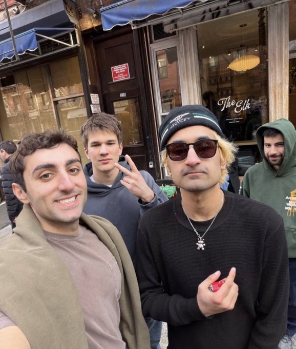 A group selfie of four young men standing outside a building; one wears a beanie and sunglasses, another wears a green hoodie, and the storefront behind them is labeled 'The Elk'.