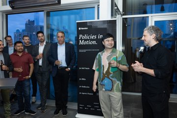 A group of people gathered at a Tokenize This NYC event standee displaying "Policies in Motion" with a speaker addressing the crowd, capturing the networking and discussion energy described in the caption about dinner with Redstone at Tokenize This NYC.