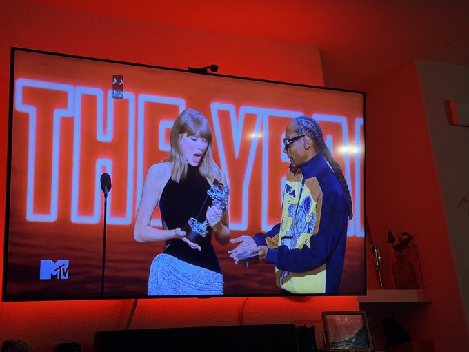 A TV screen displays a scene from The Eras Tour on MTV, showing Taylor Swift in a black top receiving flowers from a dancer, with the large red 'THE ERAS TOUR' text in the background.