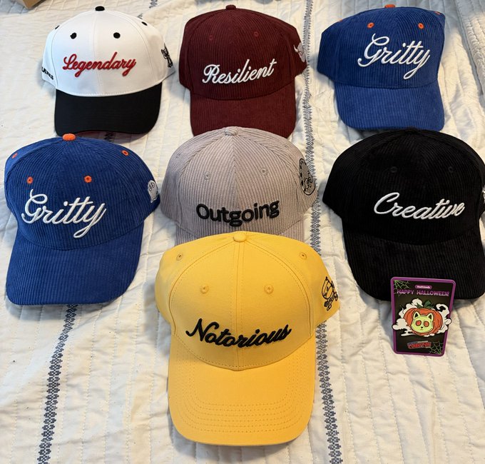 A collection of seven colorful VeeFriends baseball hats displayed on a striped bedspread, each embroidered with character trait names: "Legendary" (white/black), "Resilient" (maroon), two "Gritty" hats (royal blue), "Outgoing" (gray), "Creative" (black), and "Notorious" (yellow), along with a "Jumpy McGuffin" collector card.