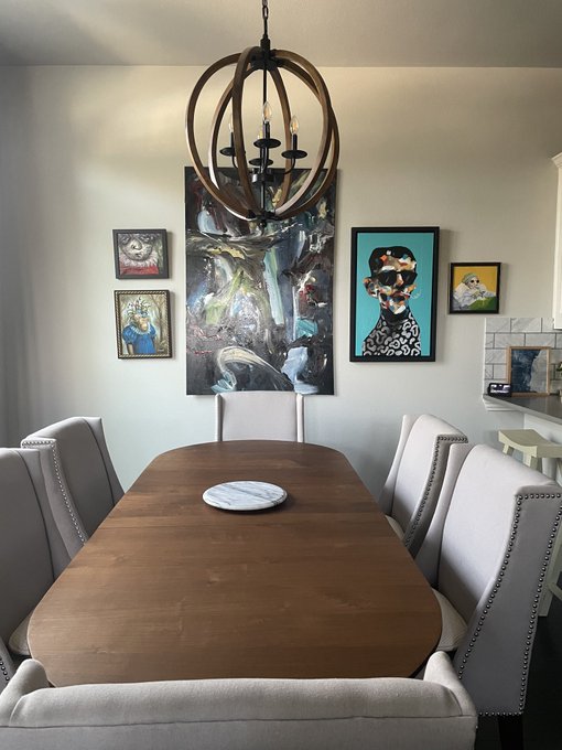 An elegant dining room featuring a long wooden table with cream-colored upholstered chairs, a wooden spherical chandelier overhead, and a gallery wall displaying various framed artworks including portraits and landscape paintings.