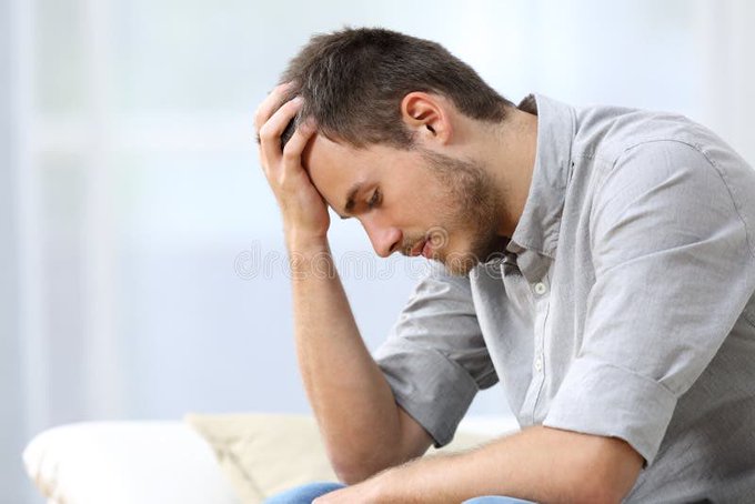 A man in a light gray shirt sits with his head in his hand, appearing stressed or deep in thought.