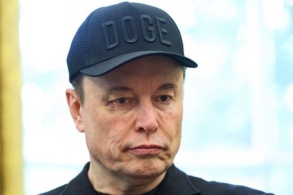 A close-up portrait of Elon Musk wearing a black cap with 'DOGE' embossed on it, looking downward with a serious expression.