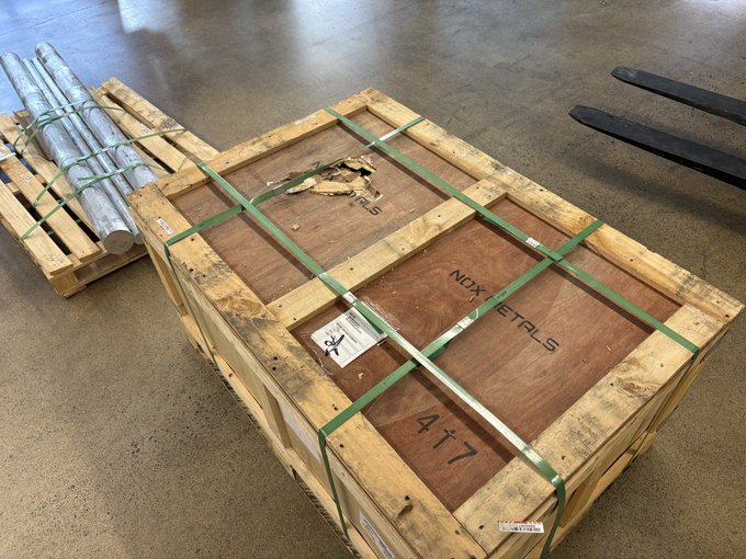 A wooden shipping crate secured with green metal strapping on a concrete warehouse floor, marked with 'NDX METALS', the number '477', and other shipping labels, sitting next to a pallet of metal pipes.