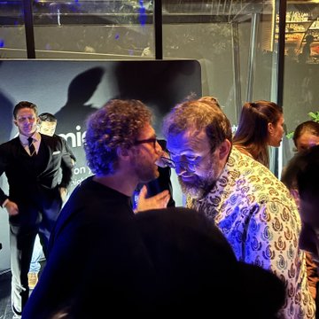 Two men in conversation at what appears to be a premiere or event, with one in a black shirt and glasses speaking closely to an older man in a patterned shirt, while another man in a suit stands in the background.