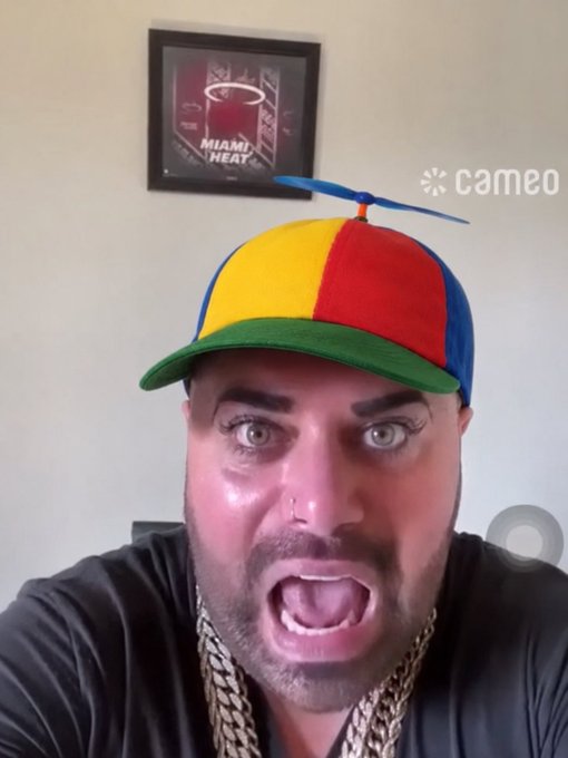 A selfie-style video still from Cameo showing a man wearing a colorful propeller hat and gold chain, with a Miami Heat framed picture on the wall behind him.