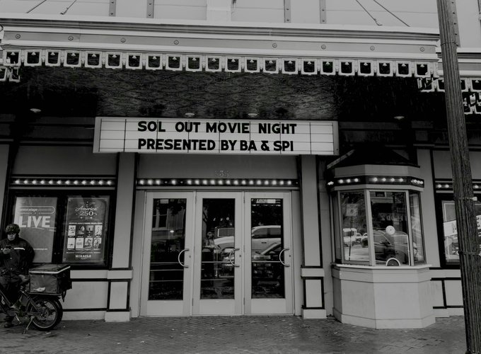A black and white photo of a theater marquee that reads 'SOL OUT MOVIE NIGHT PRESENTED BY BA & SPI' above the entrance doors, with a person standing by a bicycle on the left.
