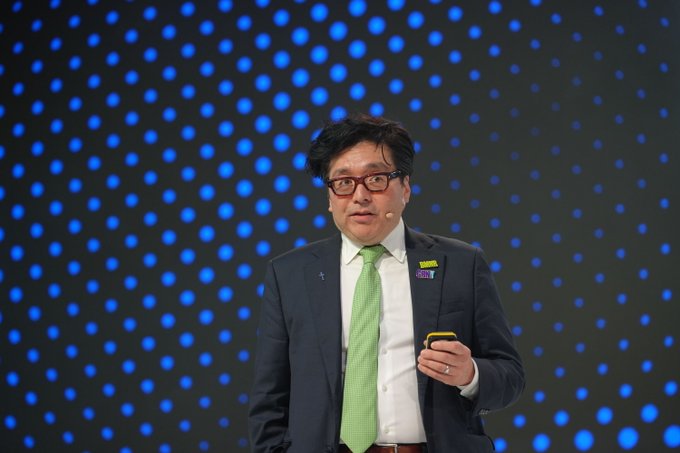 A man wearing glasses, a dark suit, white shirt, and green tie stands on a stage with a blue polka dot backdrop, holding a presentation clicker and wearing a microphone headset.