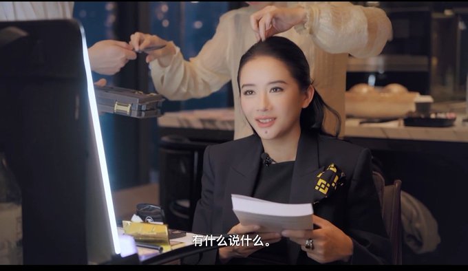 A woman in a black blazer is seated at a vanity mirror, speaking while holding a piece of paper, with Chinese subtitles displayed at the bottom of the screen.