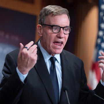 The image shows a man wearing glasses, a suit jacket, and a tie standing behind a podium with his hands outstretched as if he is giving a speech or making an announcement. The caption of the image also mentions that "he's talking about bitcoin" which refers back to the statement in the original caption. This suggests that there may be some connection between the man's words and the topic of discussion, possibly regarding Bitcoin or cryptocurrency.