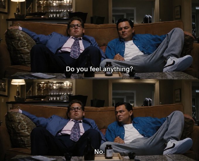 The image shows two men sitting on a couch, with one man wearing glasses and holding a beer bottle while the other man wears a suit and tie. The text above them reads "Do you feel anything?" which seems unrelated to their current situation. The main focus of the image is the two men's attire and the casual setting they are in.