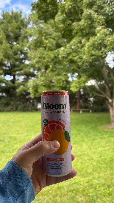 A hand holds up a can of 'Bloom' sparkling energy drink in juicy orange flavor against a backdrop of green trees and grass.