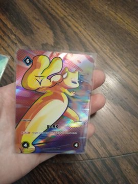 A hand holds a holographic Pokemon trading card featuring a Charmander-like creature labeled 'Rark'.